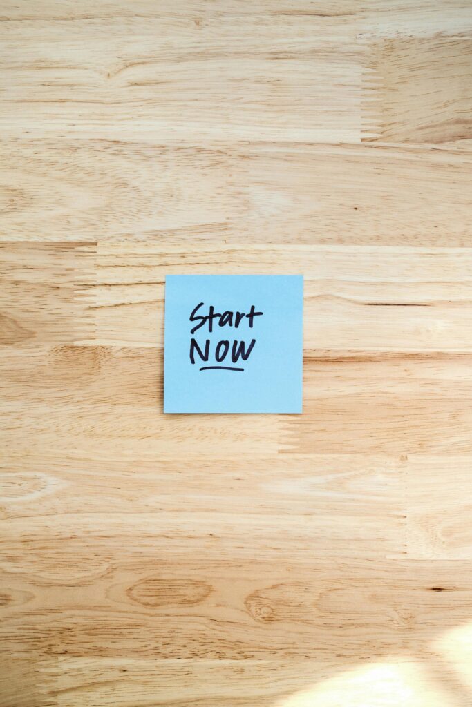 Blue Post-It note with 'Start Now' on light wooden background, inspiring action.